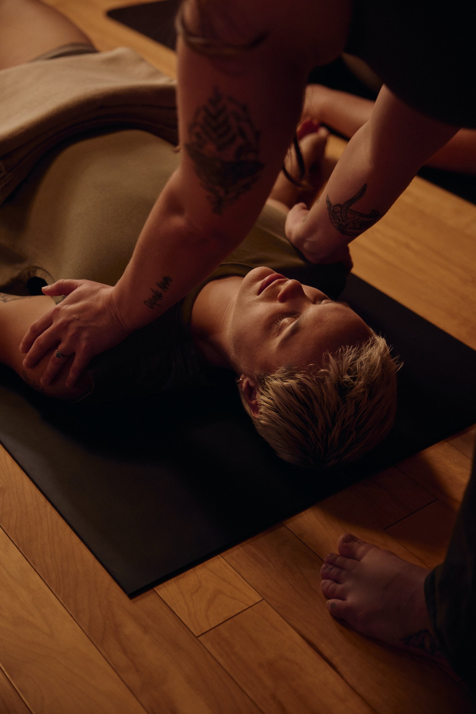 Movement instructor assists a person in a yoga pose on a black mat in warm indoor lighting