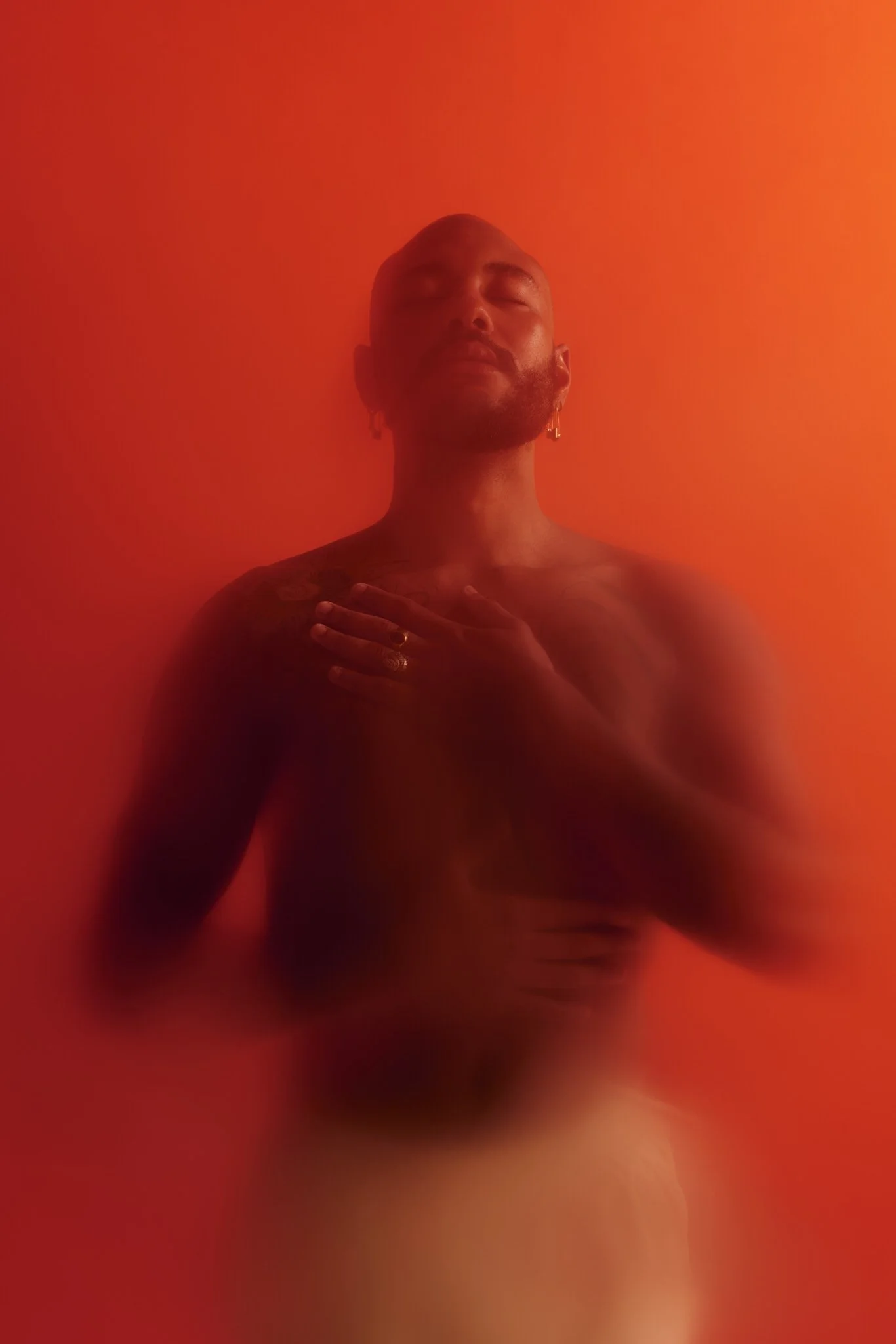 Bearded bald man with closed eyes and hands on his chest, photographed in dramatic red-orange lighting.