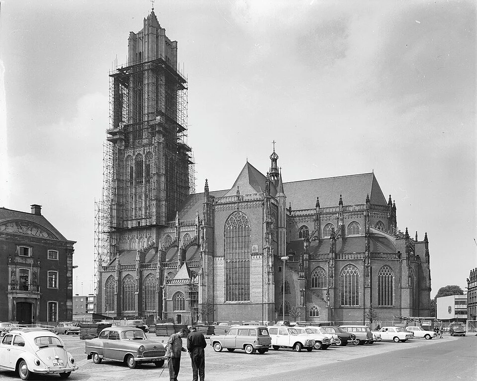 Een zwarte en witte foto van een gotische kathedraal met een toren onder constructie, omringd door oude gebouwen en een parkeerplaats met vintage auto's.