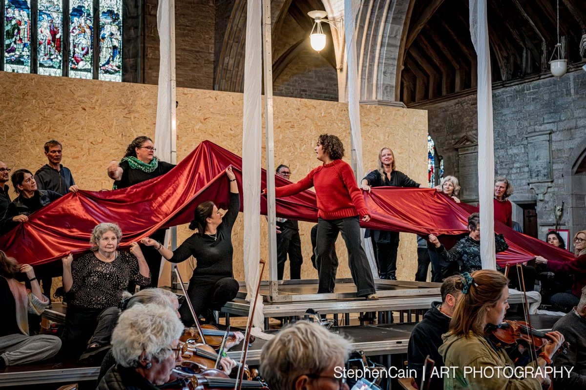 People performing on stage with a large red fabric in a church or cathedral setting, surrounded by musicians playing string instruments, stained glass windows in the background.
