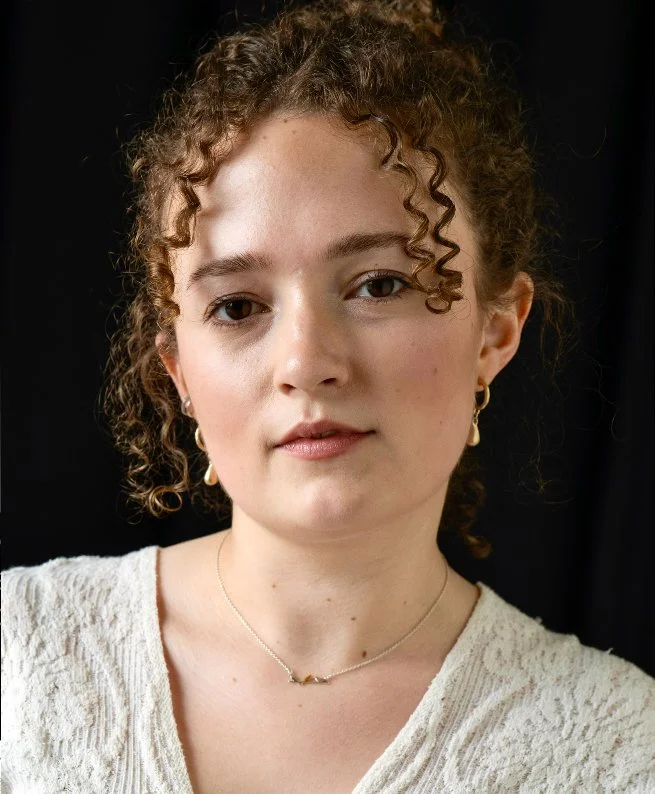 A young woman with curly light brown hair, wearing gold earrings and a delicate necklace, posing in front of a black background.