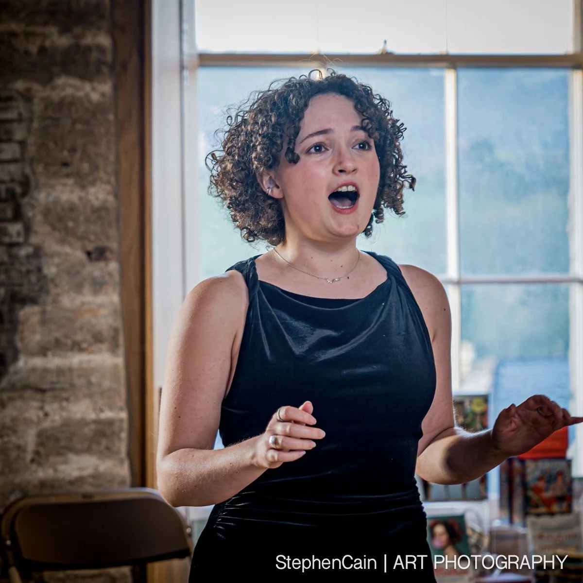 A woman with curly hair singing or speaking passionately indoors near a large window with natural light.