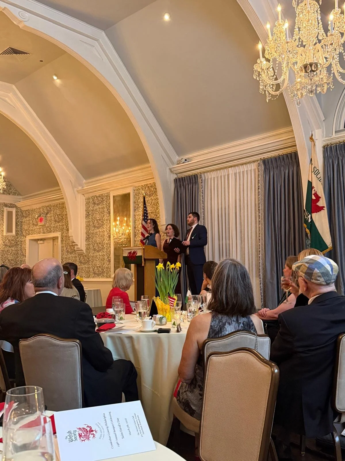 A formal event in an elegant room with chandeliers, curtains, and floral wallpaper. Three speakers are at a podium, with an American flag and a California state flag behind them. Attendees are seated at round tables, listening to the speakers.