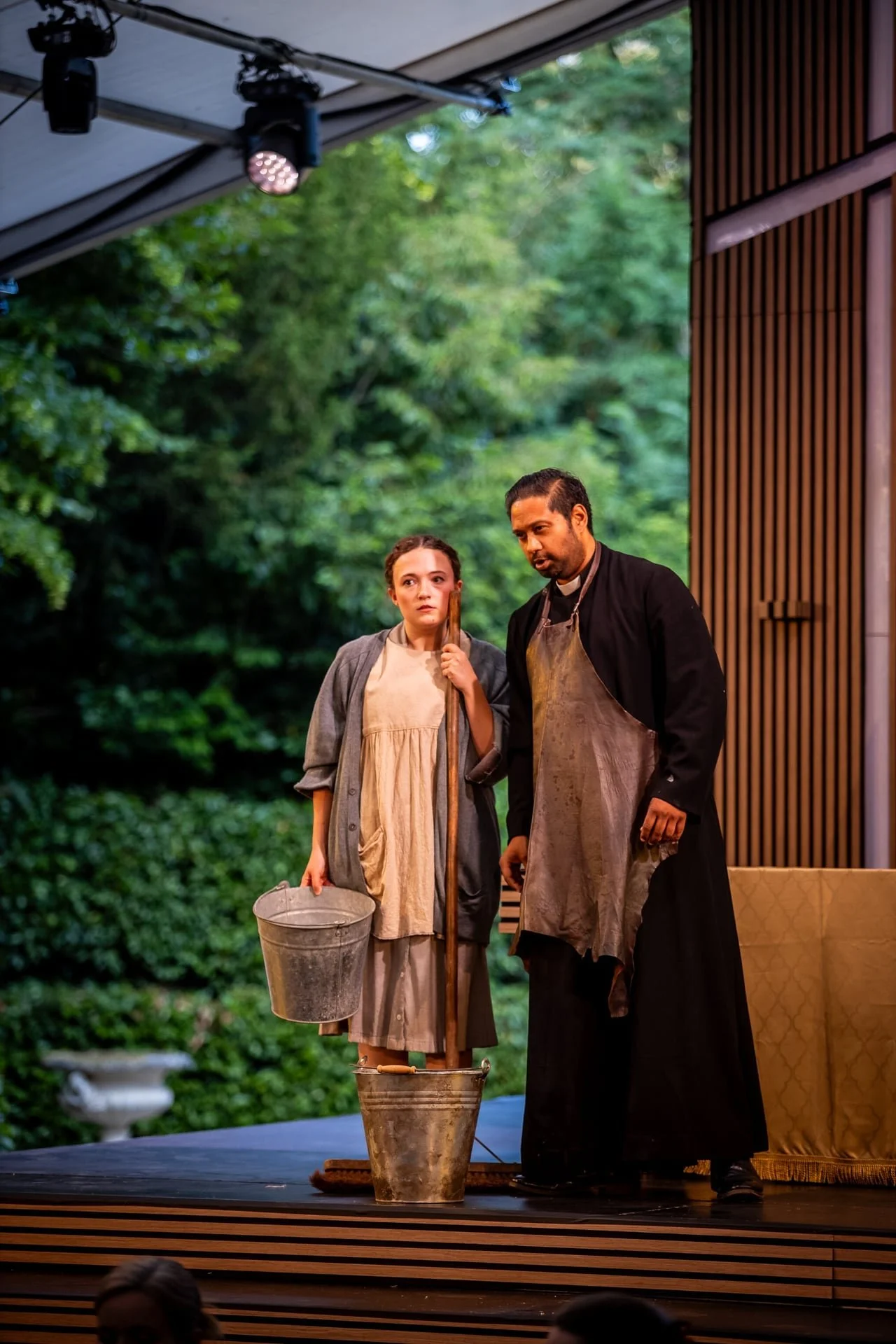 Two actors performing on stage, a woman and a man, with a lush green outdoor background. The woman holds a bucket, and the man, dressed as a priest, wears an apron. They are engaged in a scene, with focused expressions.
