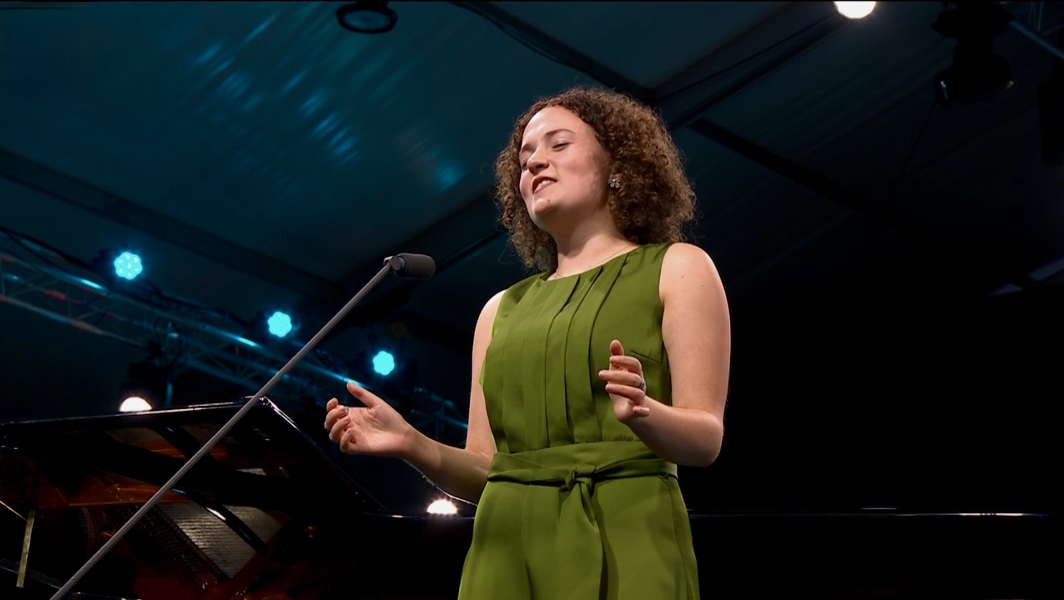 Woman with curly hair in a green dress performing at a music event on stage with blue lights.