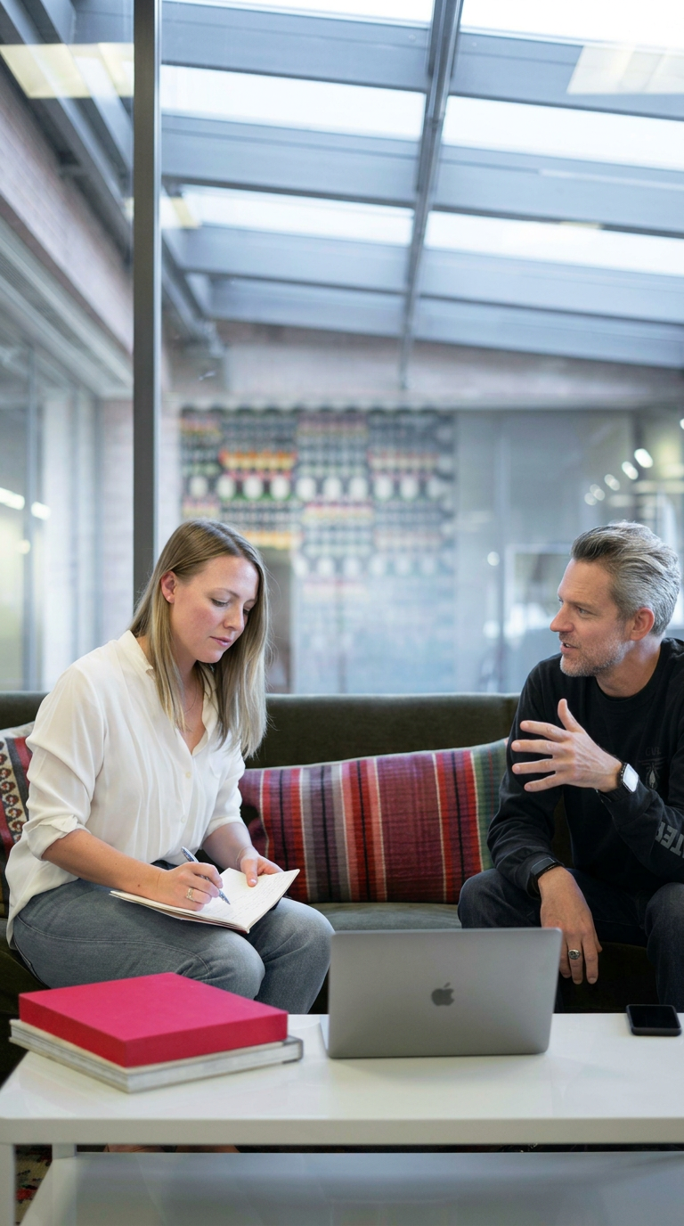 A woman and a man having a discussion in a modern office lounge area, with a laptop, notebooks, and books on a white table.