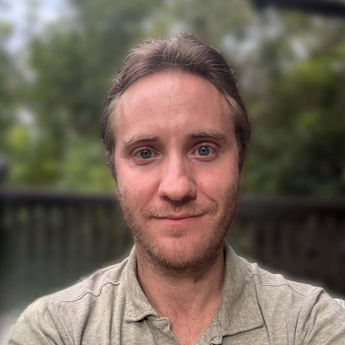 A man with light brown hair and a beard, wearing a beige shirt, standing outdoors with blurred green trees in the background.