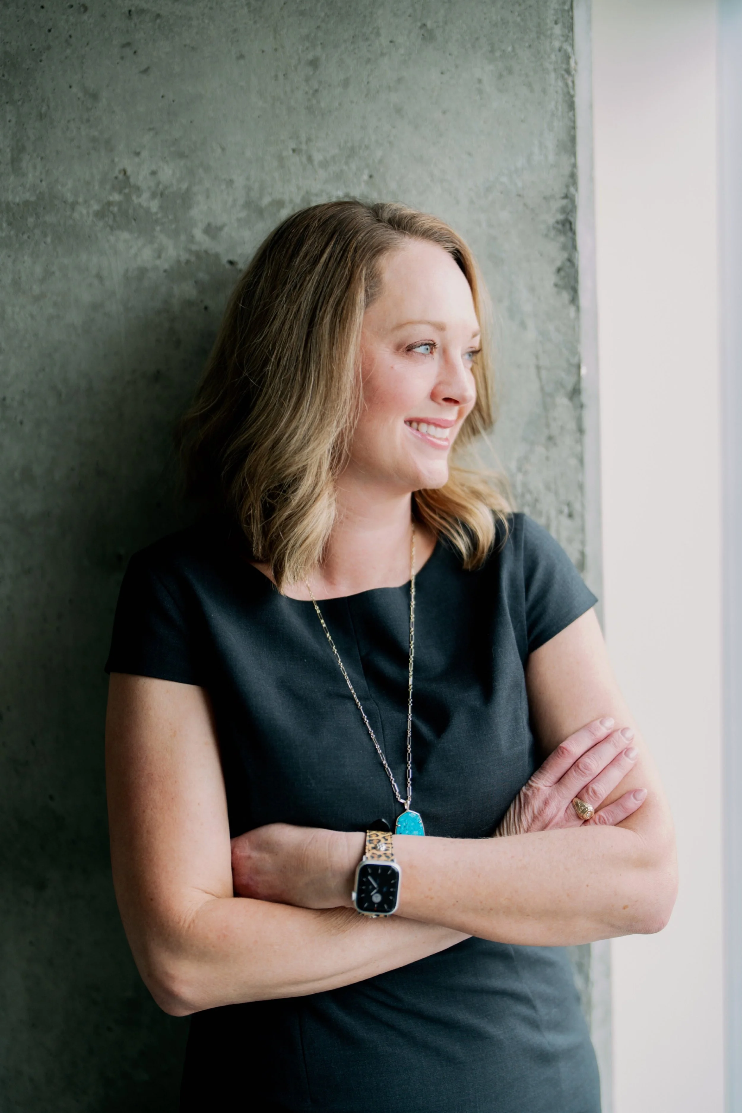 A woman with light brown hair, wearing a black short-sleeved top, jewelry including a necklace and watch, standing with arms crossed, smiling, and looking out a window, with a textured gray wall behind her.