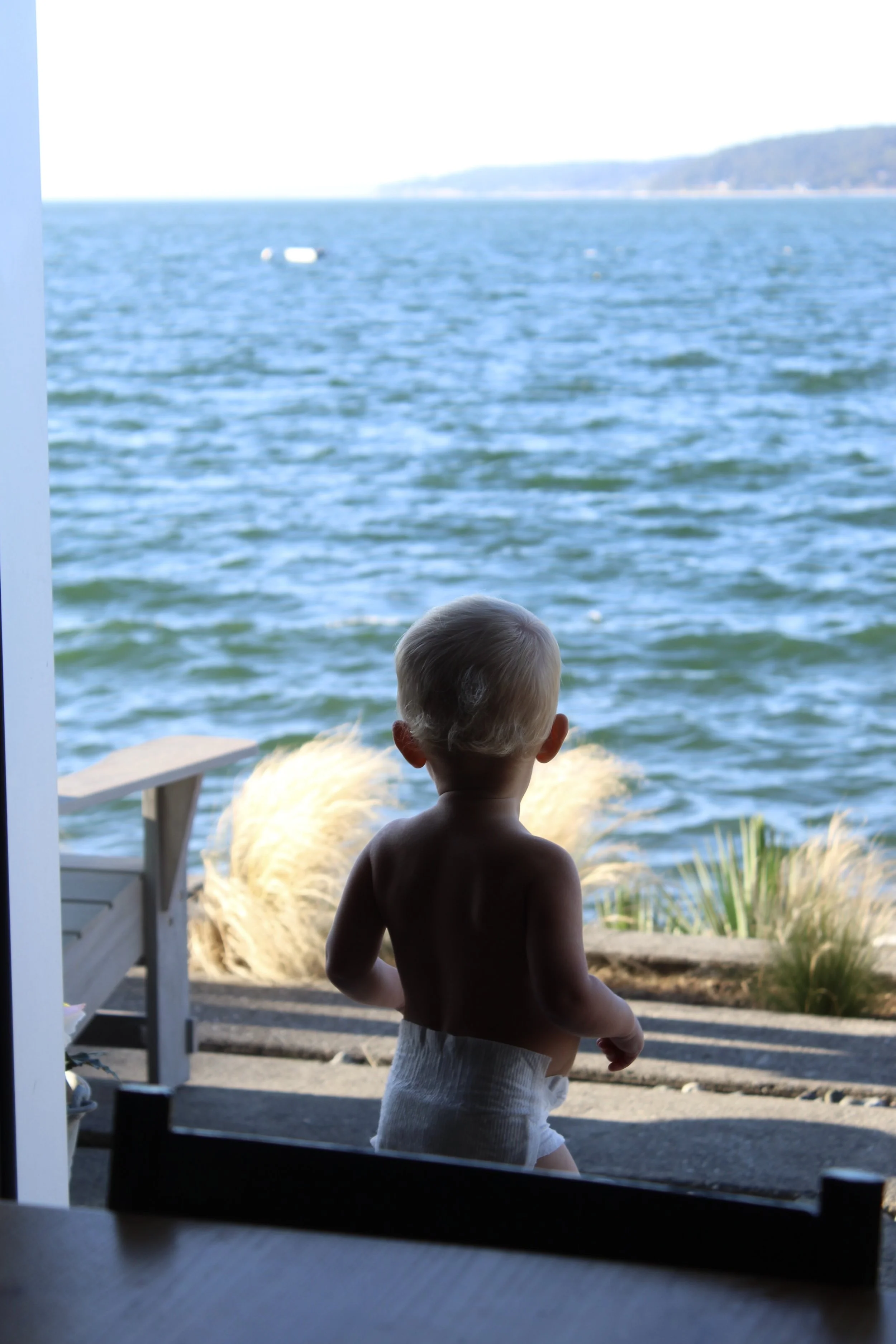 A young child with short light-colored hair and no shirt stands on a porch overlooking a body of water with hills in the distance, during daytime.