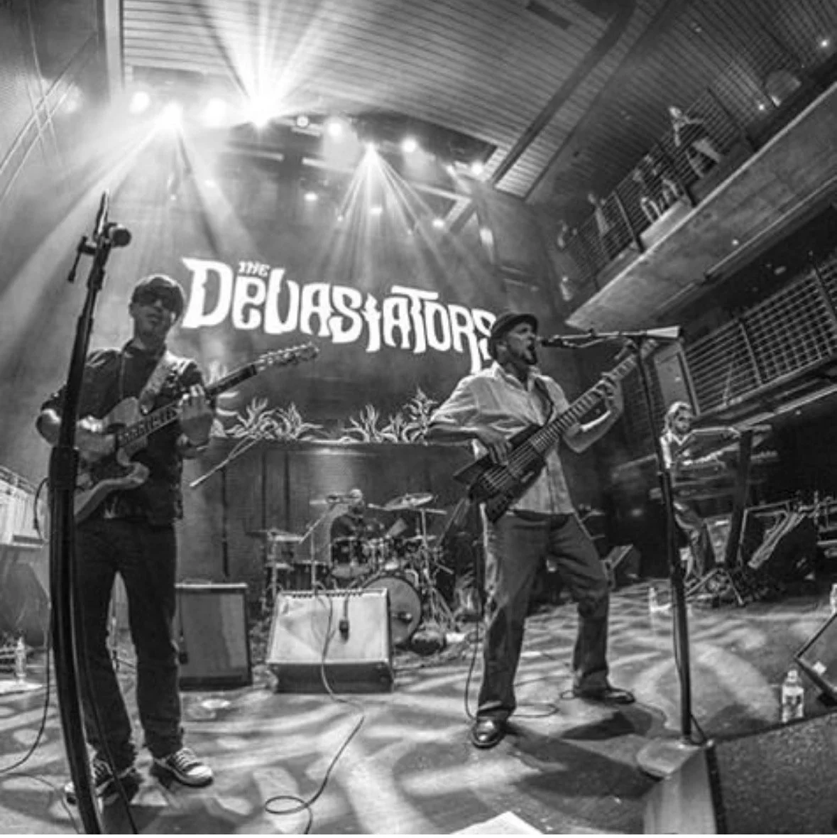 A band performing live on stage with a backdrop reading 'The Deviators.' The band members are playing guitars and drums, with stage lighting overhead and audience members visible on an upper balcony.