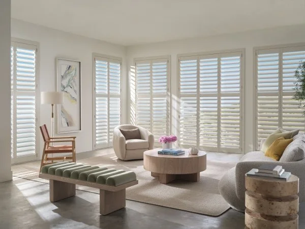 Bright living room with large windows and white plantation shutters, modern beige armchair, curved gray sofa with yellow and white pillows, round wooden coffee table with pink flowers and books, upholstered bench, side table with stacked books, and a