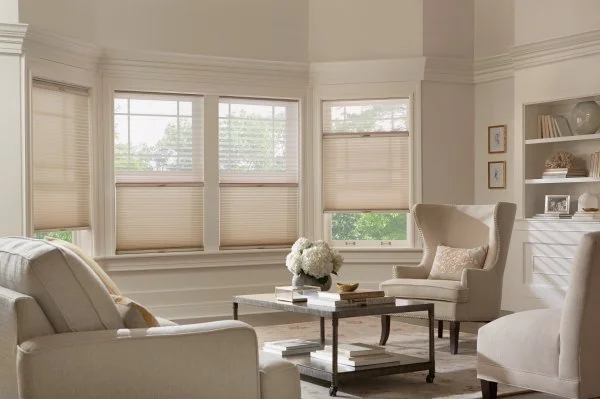 A bright living room with multiple windows, beige blinds, white walls, and cream-colored furniture, including a sofa and armchair. A coffee table with books and flowers is in the center, and built-in shelves with decorative items are on the wall.