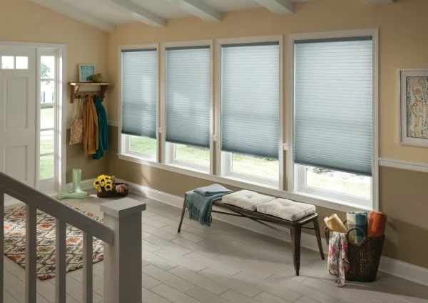 Sunny porch with four large windows fitted with blue blinds, a small wooden bench with cushions, a coat rack with jackets, a decorative vase with sunflowers, a colorful rug, and a basket containing fabric rolls.