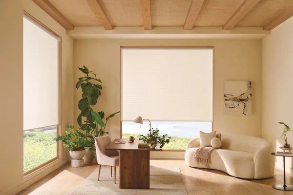 Living room with large windows, wooden ceiling, beige sofa, potted plants, wooden table, pink chair, and neutral decor.