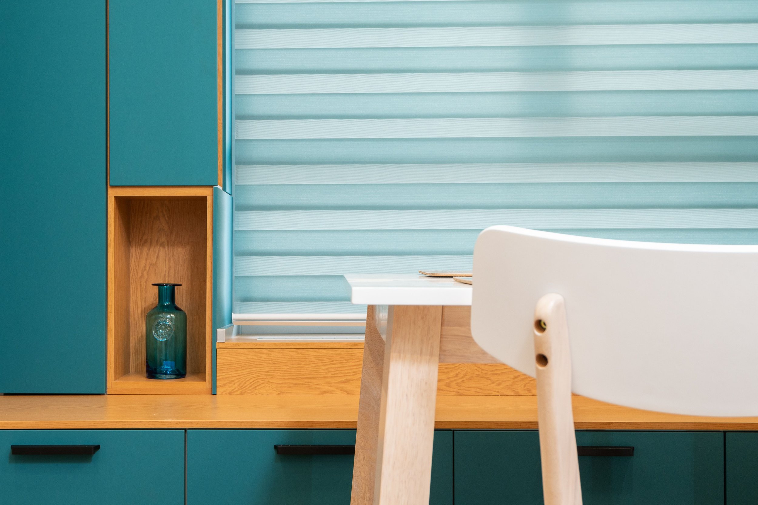 Modern kitchen with teal cabinets, a wooden open shelf with a blue glass vase, and a striped window shade. Part of a white chair and a wooden table are visible in the foreground.