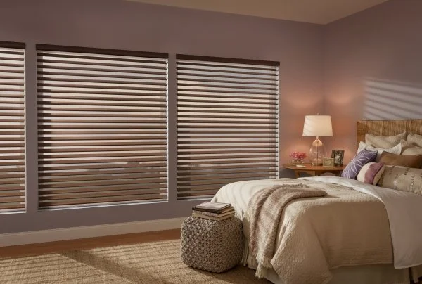 Bedroom with three striped window blinds, a bed with pillows and a blanket, a nightstand with a lamp, picture frames, and flowers.