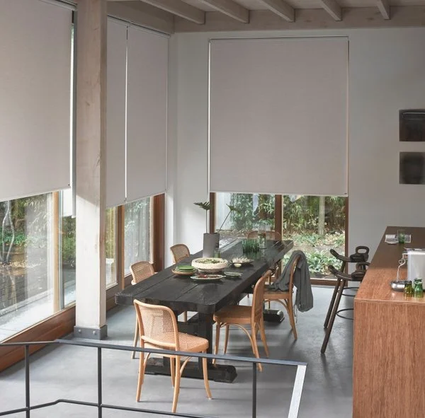 Dining area with a black wooden table, six wicker and wood chairs, set with plates and bowls. Large windows with white blinds and a view of outdoor greenery.