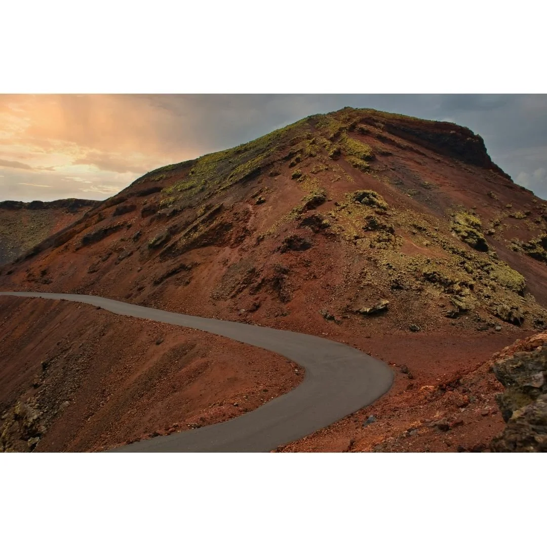 Eine kurvige Straße führt an einem Kargelandschaft mit rotbraunem Boden und einem Hügel mit grüner Vegetation vorbei, bei Sonnenuntergang.