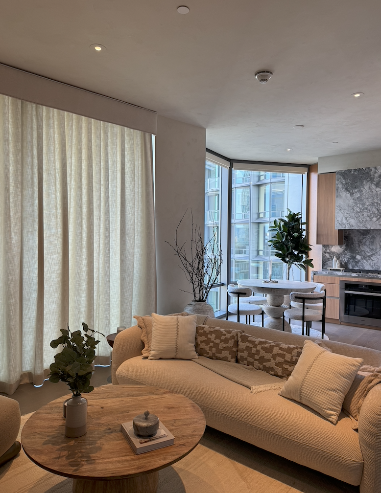 Modern living room with beige sofa, round wooden coffee table, and floor-to-ceiling windows with light curtains. Dining area in the background features a round table, four chairs, and large potted plants.