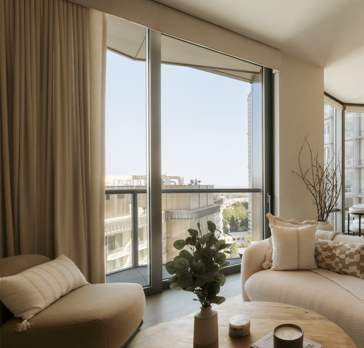 Living room with large glass sliding door leading to balcony, beige curtains, cream sofa with pillows, potted plant, and natural sunlight.
