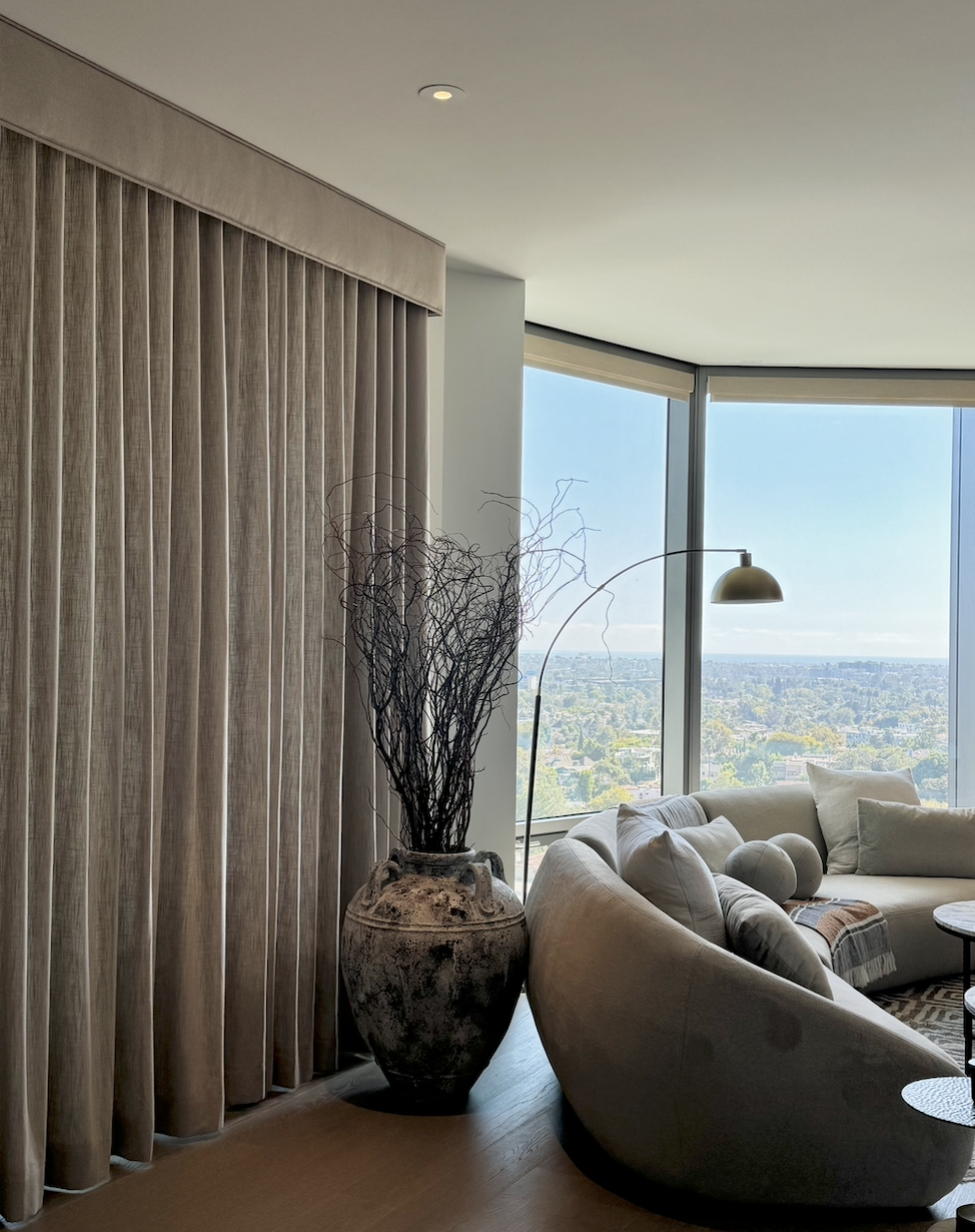 Living room with large floor-to-ceiling windows, beige curtains, a beige curved sofa with pillows, a floor lamp, a large decorative vase with dried branches, and a view of a cityscape outside