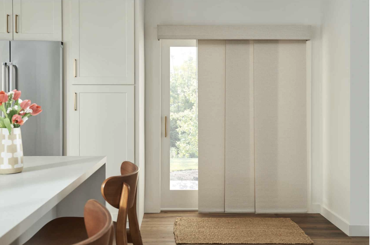 Interior view of a kitchen with white cabinets, a countertop with a vase of pink flowers, a brown chair, a refrigerator, a sliding door with beige blinds, and a woven rug on wooden floor.