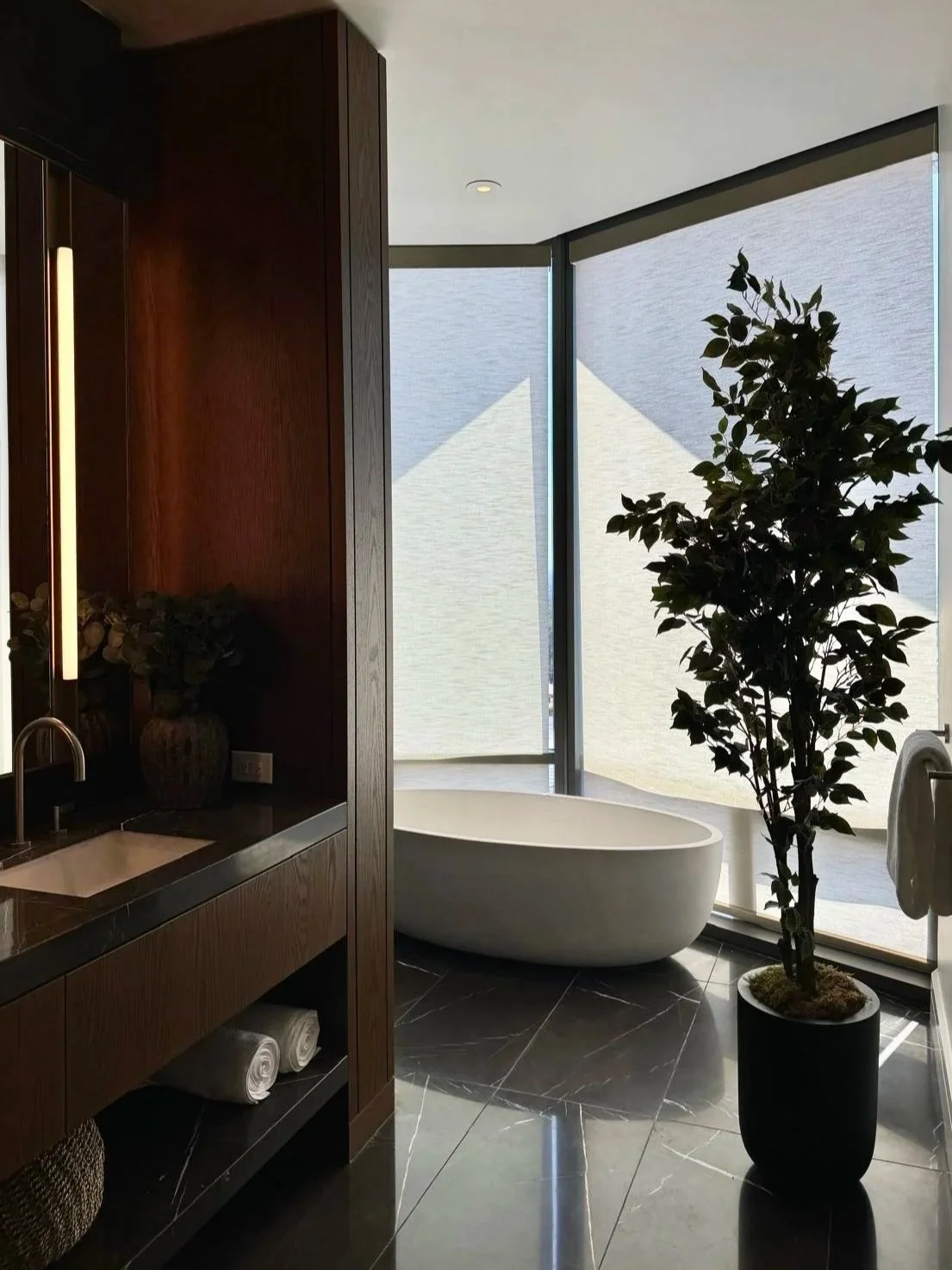 Modern bathroom with a freestanding bathtub near large windows, potted plant, dark marble floor, wooden cabinetry, and towel storage.