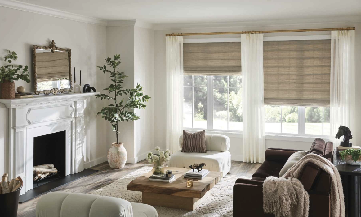 Bright living room with large windows, beige and white curtains, a white fireplace mantle, a large potted plant, and varied seating including a cream armchair, a brown sofa with a throw blanket, and a small chaise lounge with a pillow. A wooden coffe