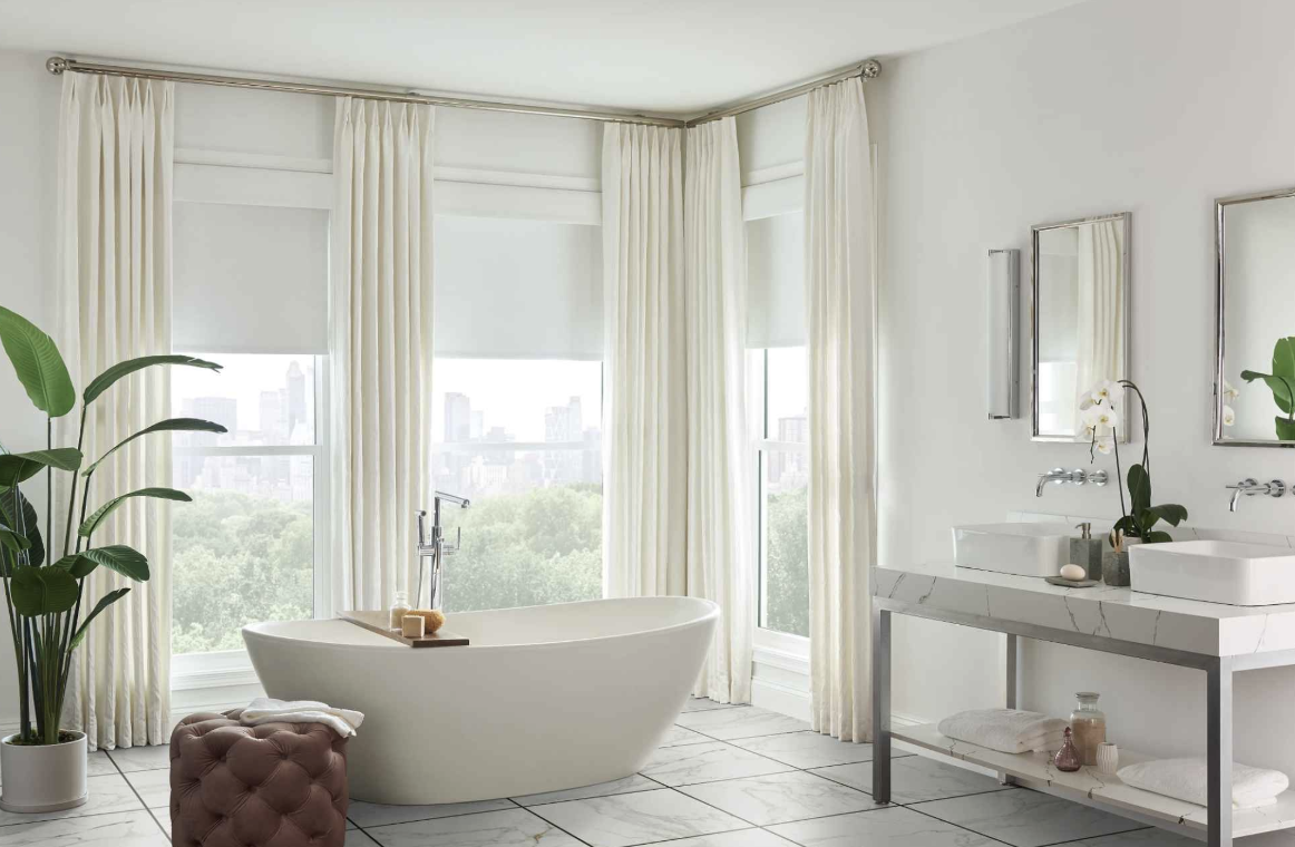 Bright, modern bathroom with a freestanding bathtub by large windows with white curtains, a double vanity with two sinks, mirrors, and potted plants.