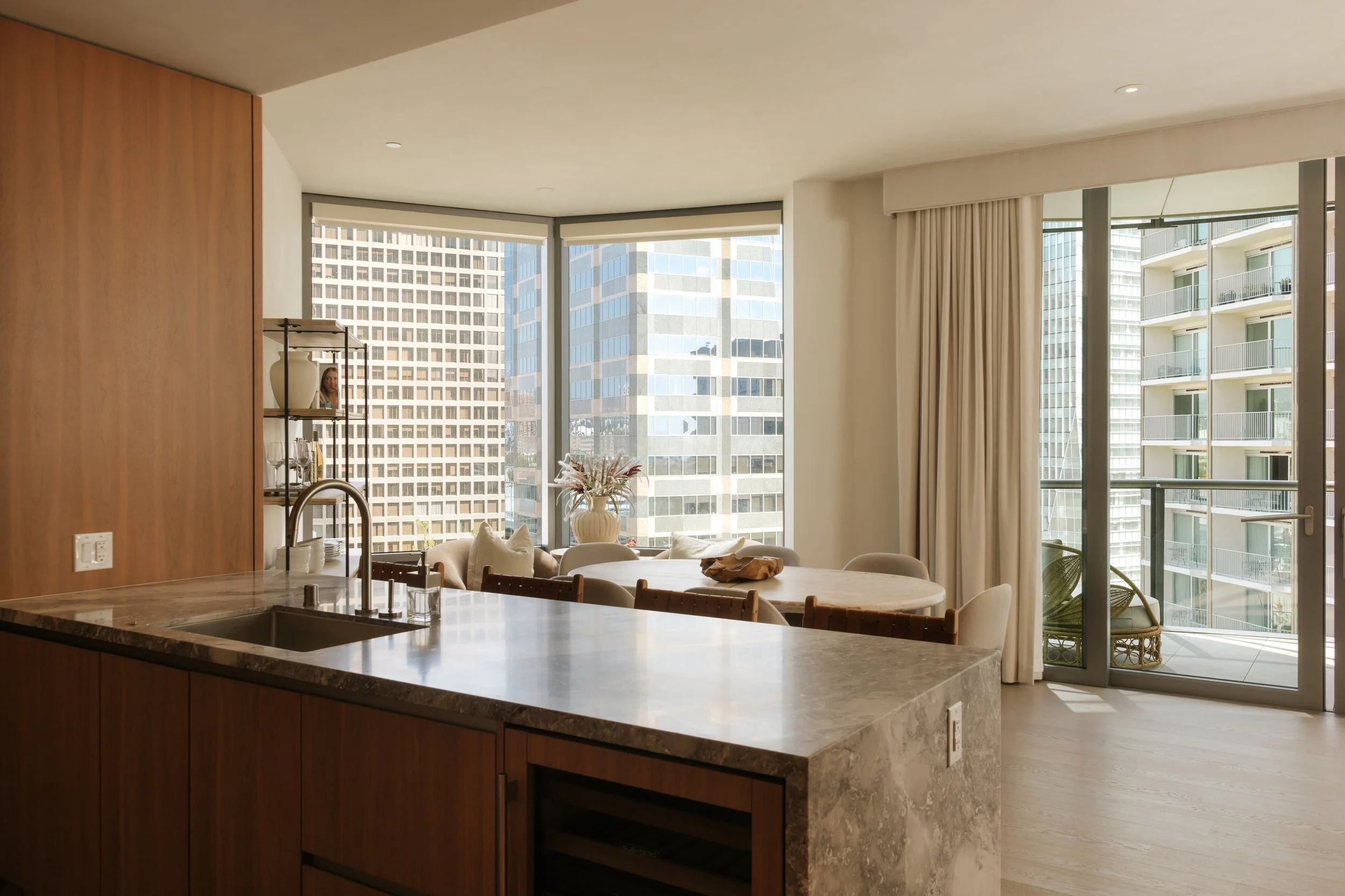 Modern apartment interior with open living and kitchen area, large windows with city views, dining table, beige curtains, and decorative items.