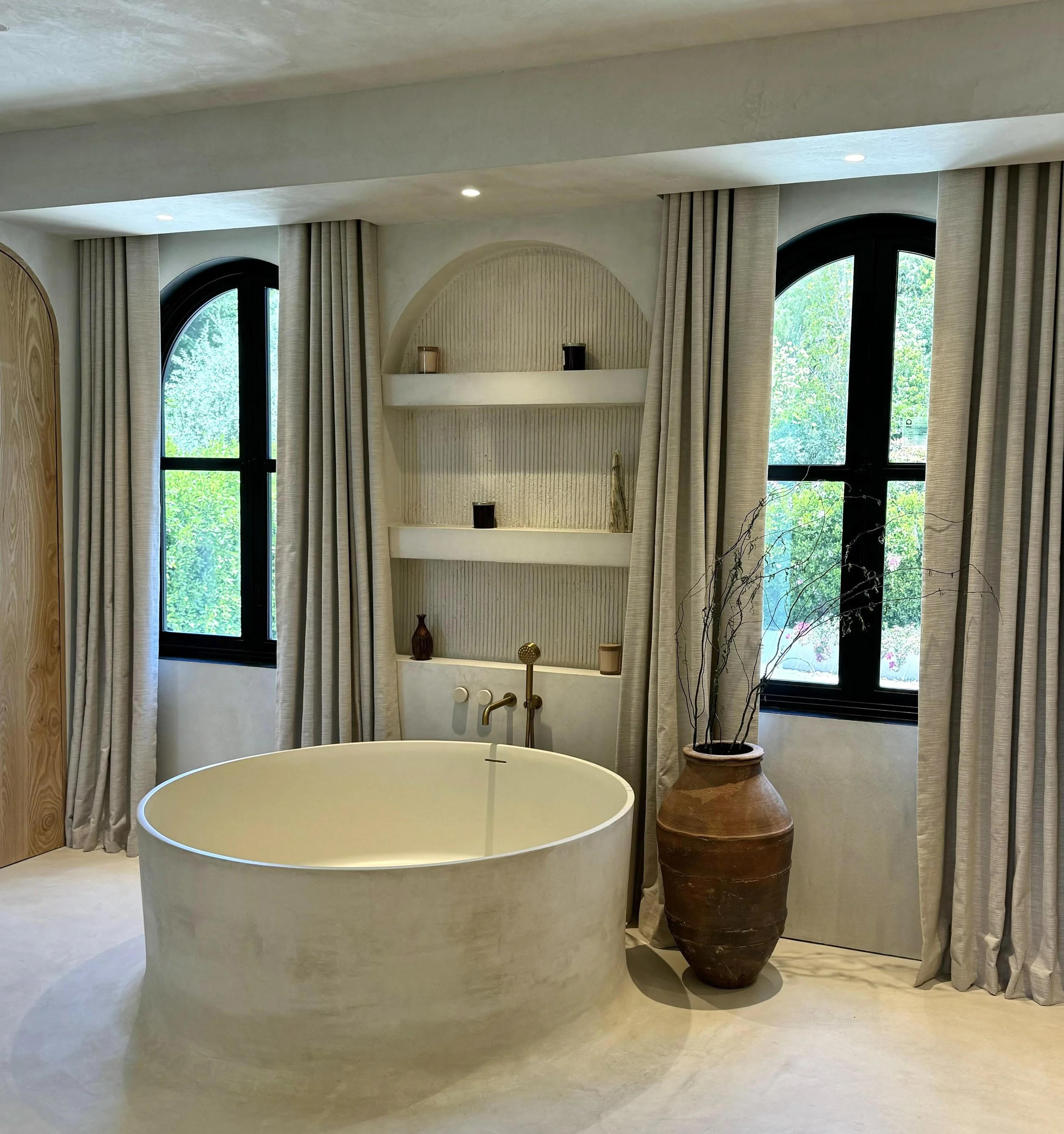 Modern bathroom features a free-standing bathtub with a circular shape, large arched windows with black frames, beige curtains, decorative vases on a built-in shelf, and a large clay pot with dried branches beside it.