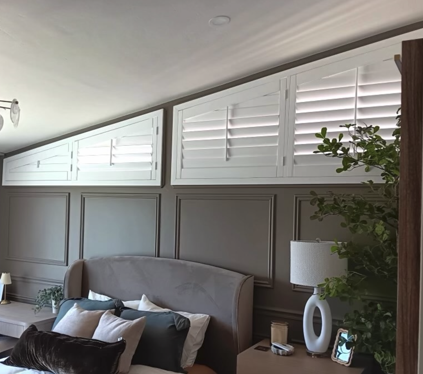A bedroom with a bed, pillows, a nightstand, a table lamp, a decorative lamp, a plant, and window shutters over the windows.