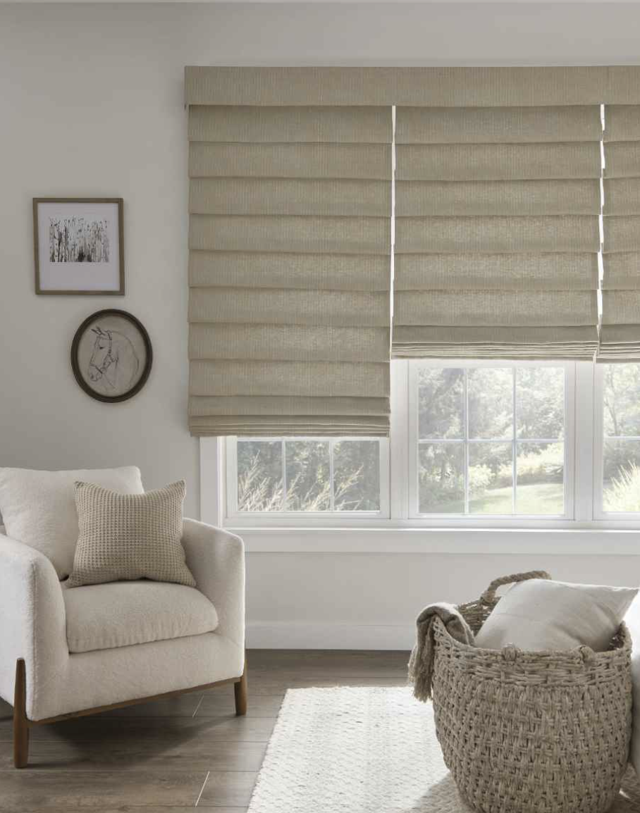 A cozy living room with beige Roman shades covering a large window, a cream upholstered armchair with a textured pillow, a woven basket with a blanket on a nearby chair, a white rug, and framed wall art including a deer sketch.