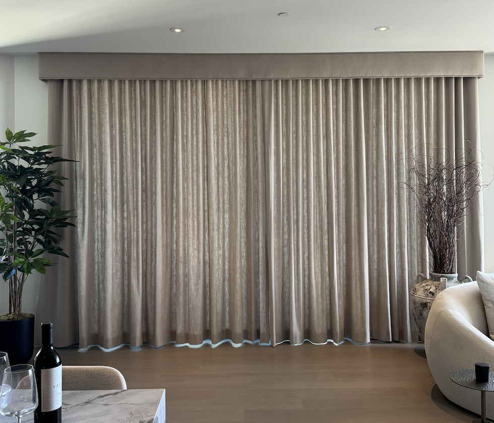 Living room with beige curtains, a green leafy plant on the left, a large ceramic vase with twigs on the right, and part of a white sofa visible.