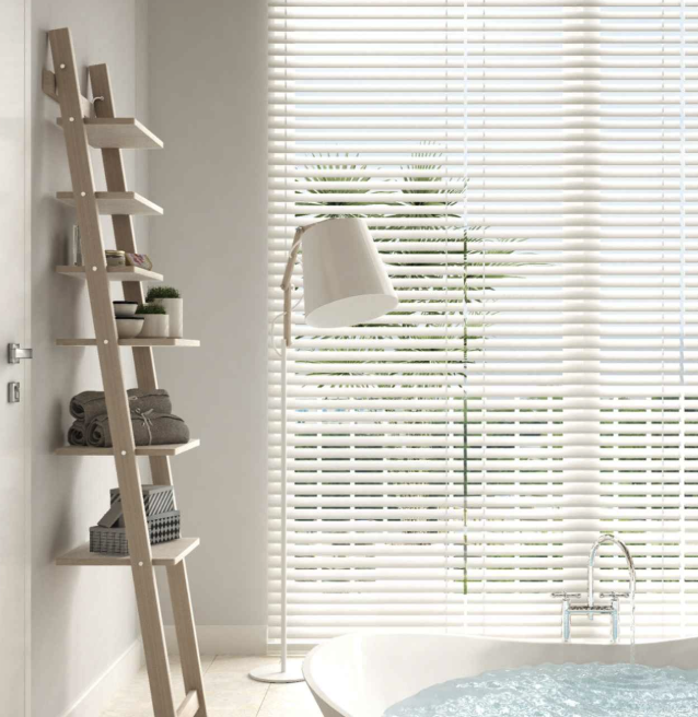 A bathroom with a large bathtub, beige ladder-style shelf on the wall filled with towels and decorative items, a floor lamp with a white shade, and large window blinds allowing natural light in.