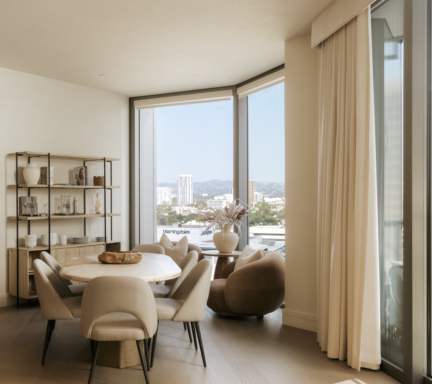 A modern dining area with a round marble table surrounded by beige chairs, a small table with a vase of pink flowers, and large windows with a city view and white curtains.