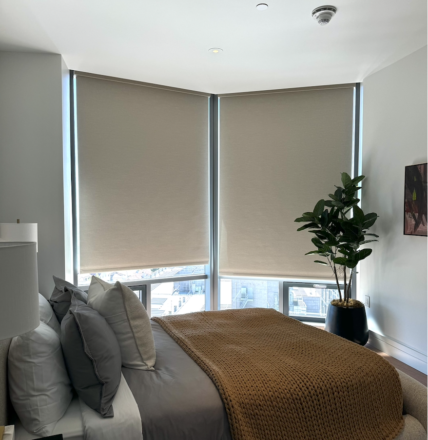 A cozy bedroom with a bed covered in white and gray linens, a chunky brown knitted blanket, and decorative pillows. Large floor-to-ceiling windows with beige blinds partially drawn, and a green potted plant near the window.