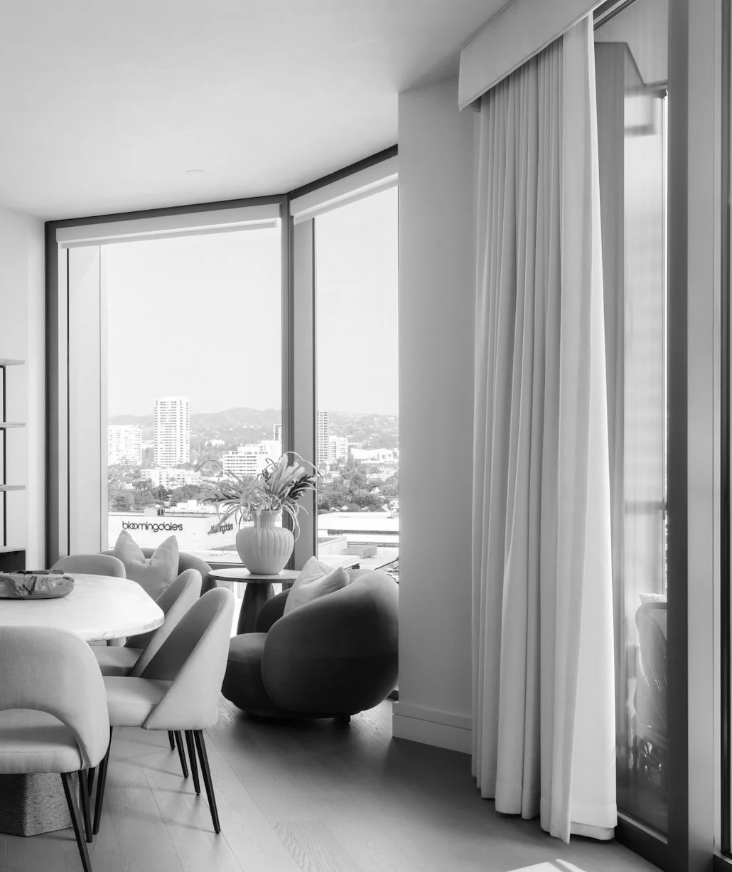 Interior view of a modern living room with large windows, white curtains, a dining table with chairs, and a cityscape outside.