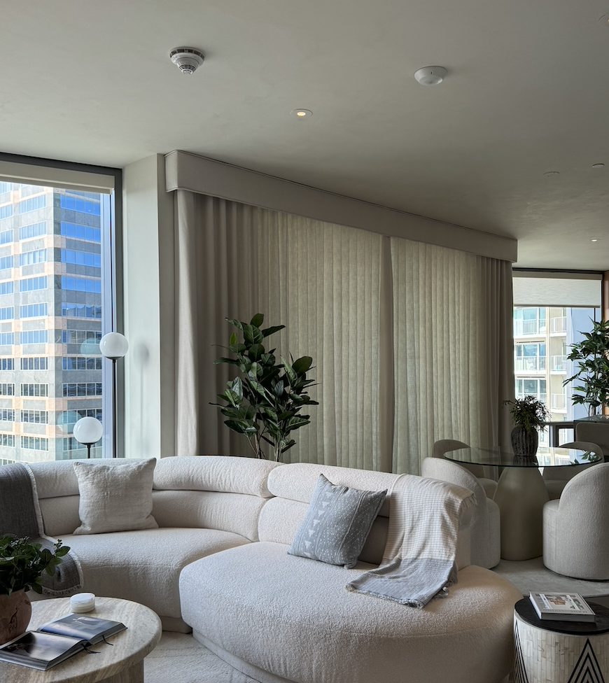 Modern living room with white curved sofa, throw pillows, and a blanket, large windows with city views, indoor plants, a round dining table with chairs, and beige curtains.