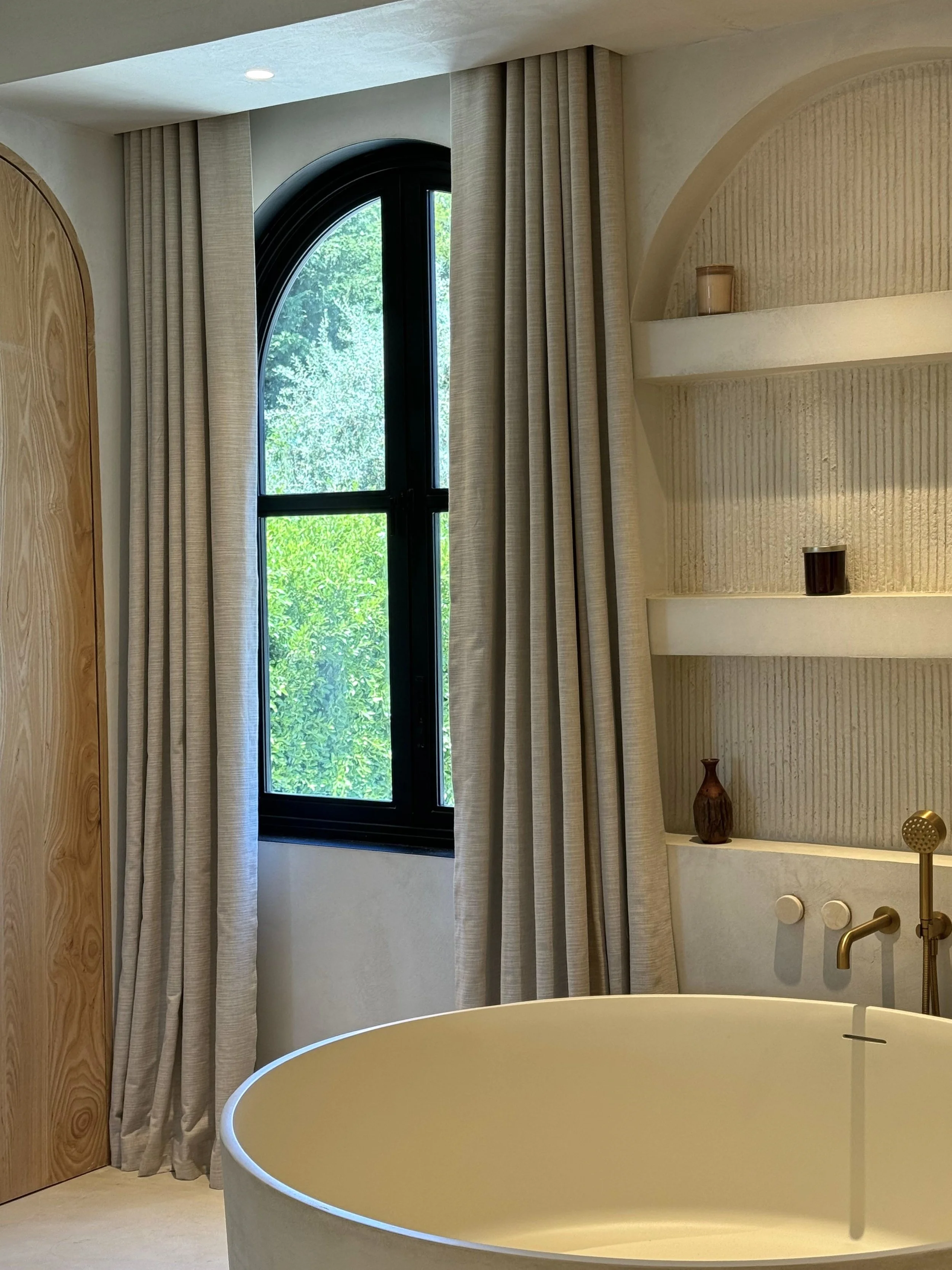 A bathroom with a curved bathtub, a large arched window with black trim, beige curtains, a textured wall with built-in shelves holding vases, and a brass faucet.