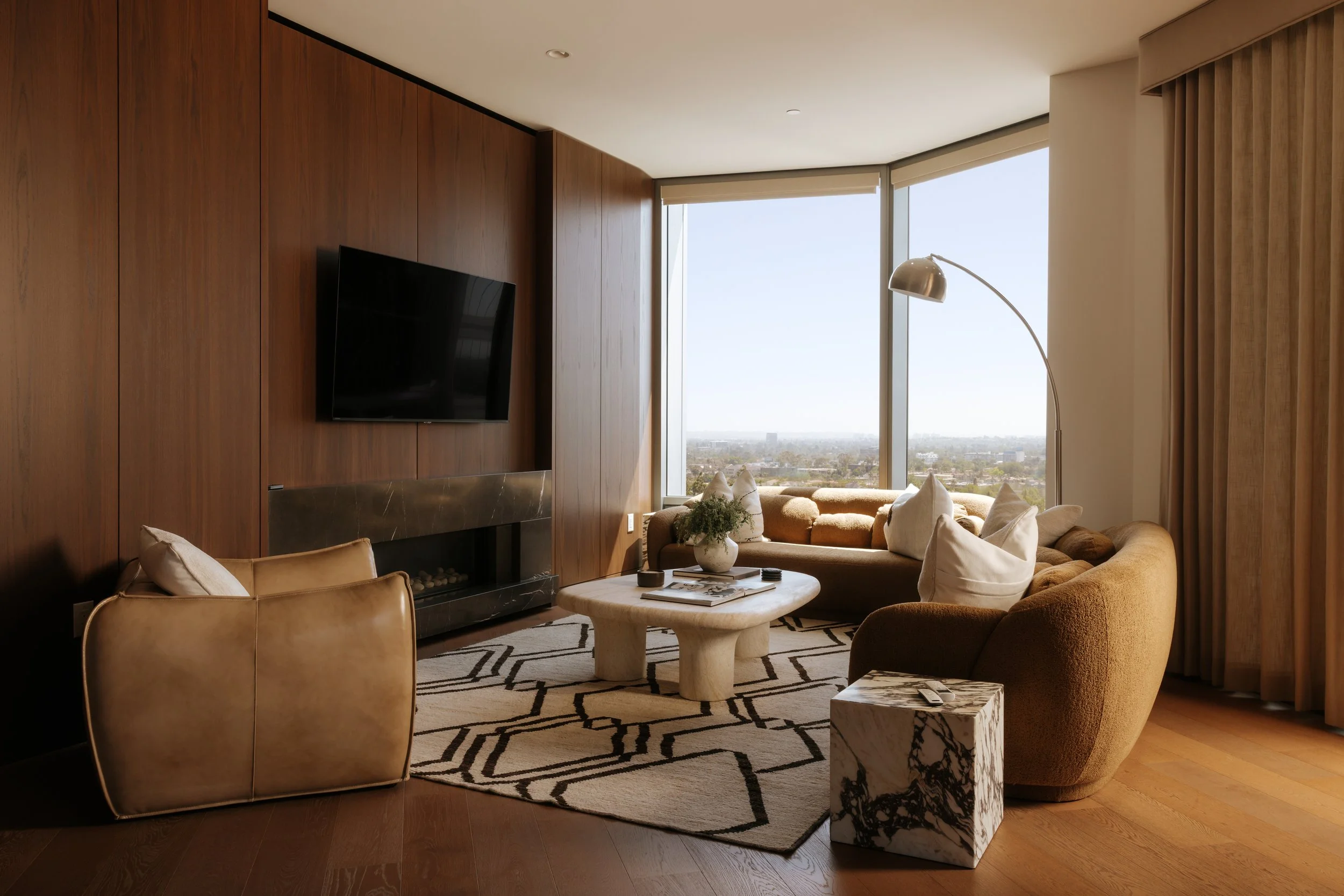 Living room with a wood-paneled wall, mounted flat-screen TV, beige armchair, curved sofa with white pillows, black-and-white geometric rug, round coffee table, marble side table, large window with city view, and a chrome arc floor lamp.