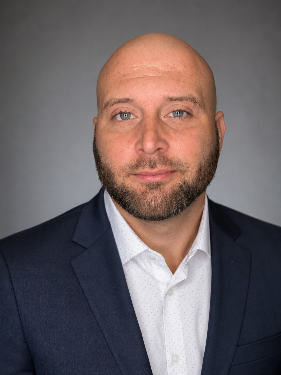 Portrait of a bald man with a beard wearing a navy blazer and white dress shirt, against a plain gray background.