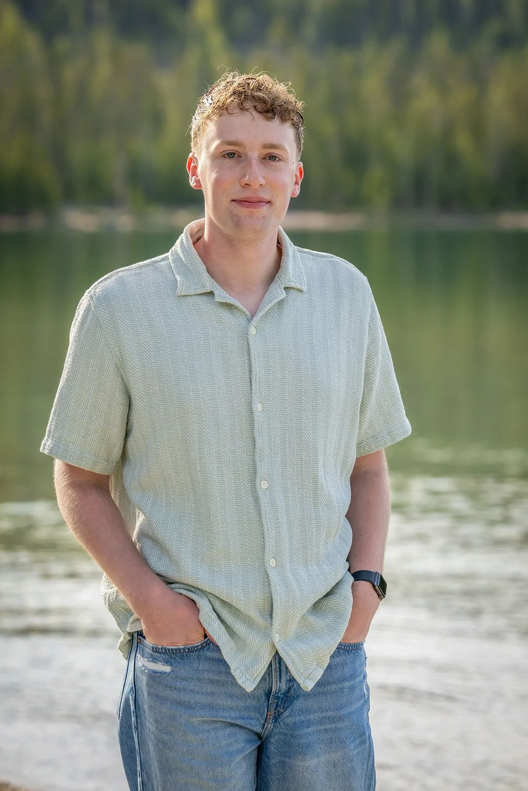 A young man with curly light brown hair and fair skin standing outdoors near a body of water, wearing a short-sleeved light-colored button-up shirt and blue jeans, with a natural background of trees and water.
