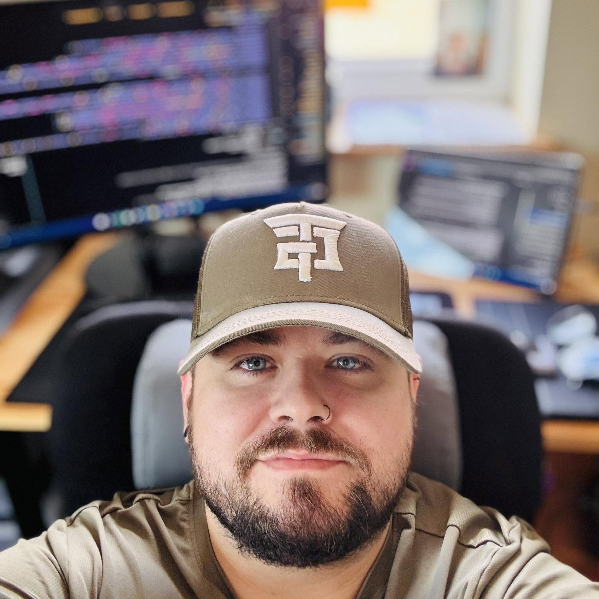 Close-up of a man with blue eyes and facial hair, wearing a beige cap with a logo, leaning forward with a blurred background of bookshelves and window.