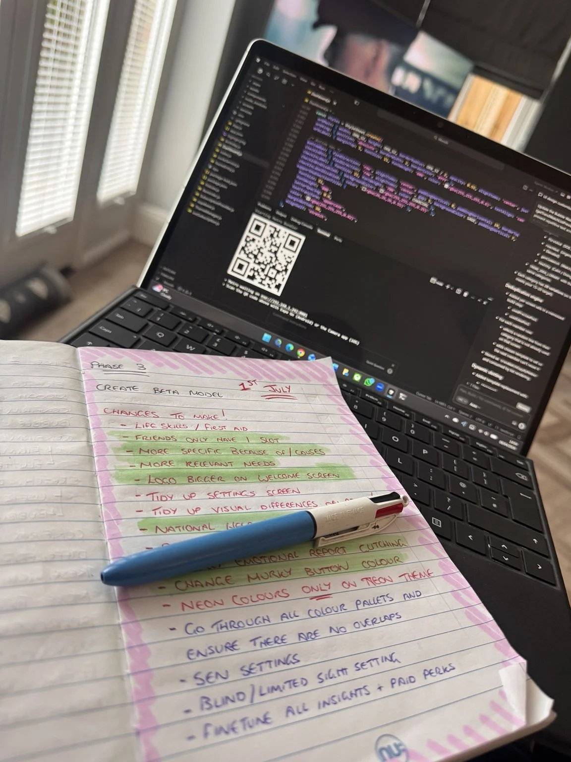 Open notebook with handwritten notes and a blue pen on top, laptop displaying a coding interface with a QR code, in a bright room with vertical blinds.