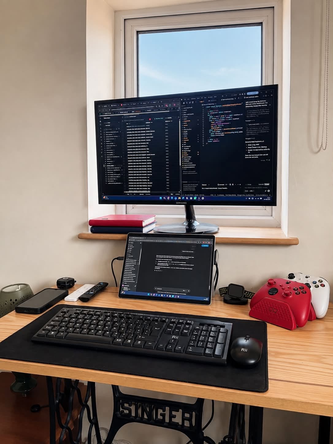 A computer workstation with a large monitor, a tablet displaying code, a keyboard, a mouse, gaming controllers, and various electronic accessories on a wooden desk by a window.