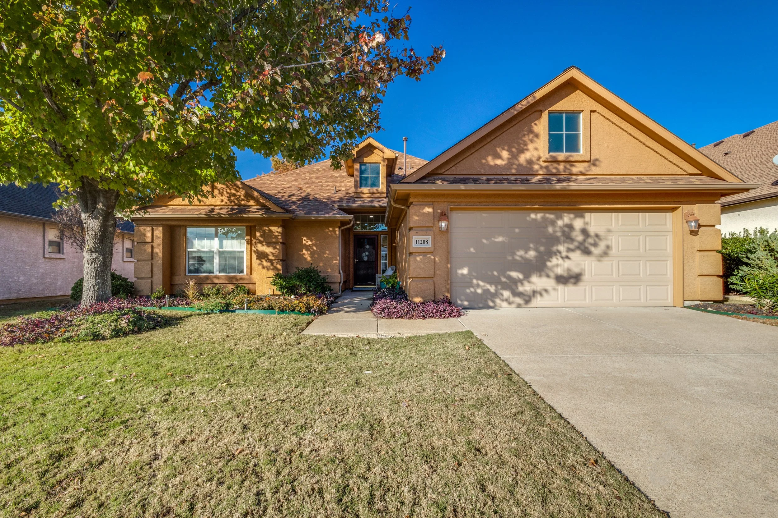 Front view of a beige house with a attached garage, a front door, small windows, and a well-maintained front yard with a tree, shrubbery, and flower beds under a clear blue sky.