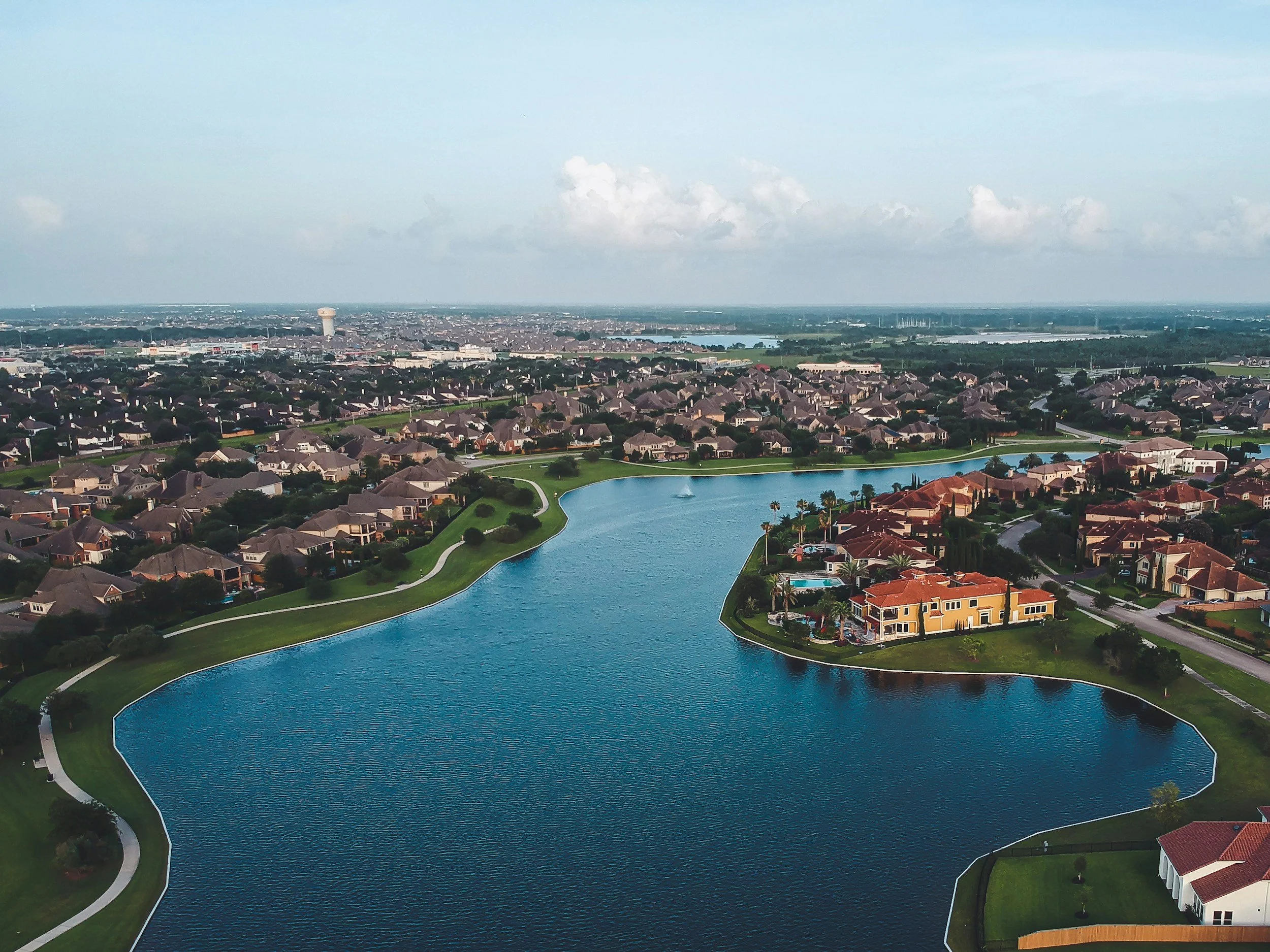 Aerial view of a suburban neighborhood with houses surrounding a large man-made lake, with a water tower and cityscape in the distant background.