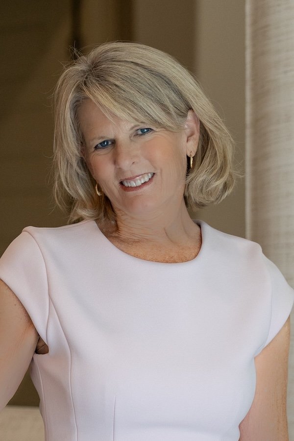 A middle-aged woman with blonde hair, blue eyes, and a friendly smile, wearing a light pink short-sleeve top and gold hoop earrings, standing indoors with a neutral background.