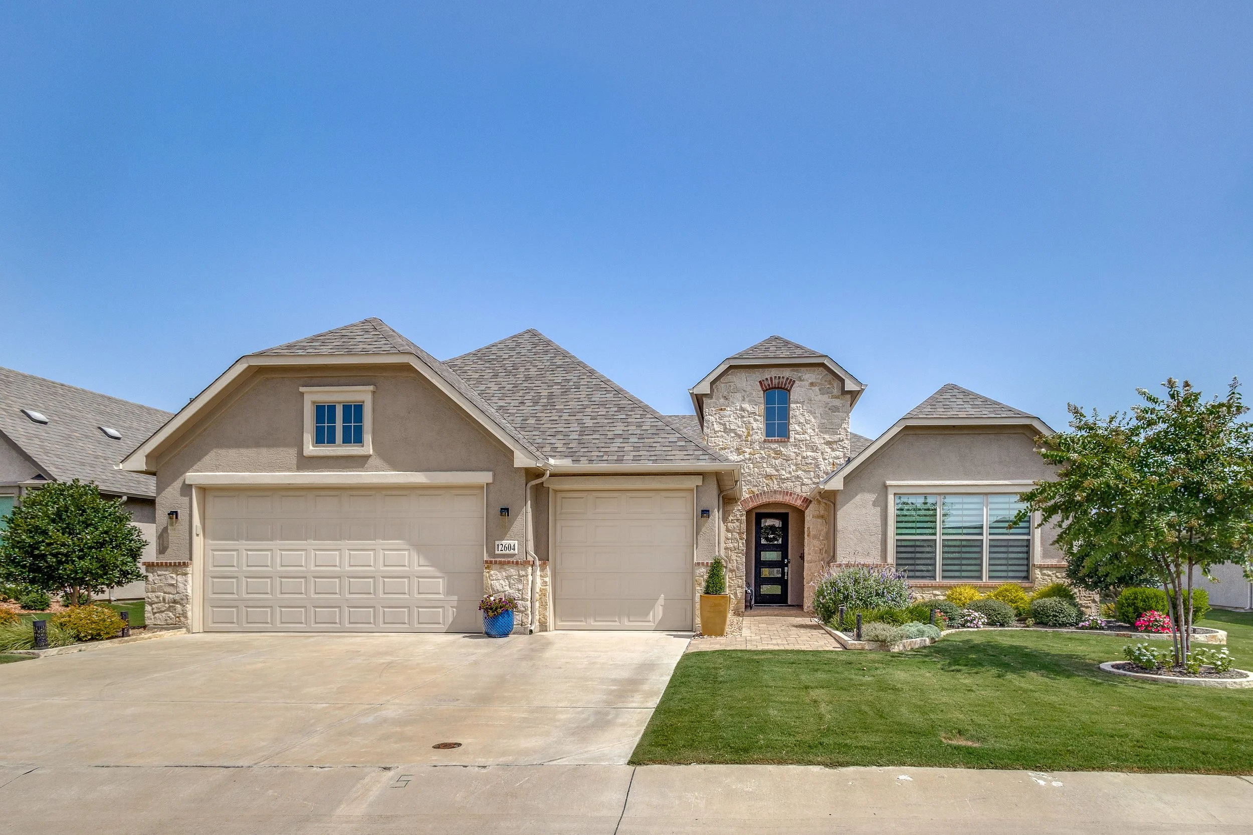 A modern suburban house with a beige exterior, two garage doors, and a front entrance with a black door. The house has a stone accent wall and some windows, including a large window with blinds. The front yard has green grass, trees, and colorful flo
